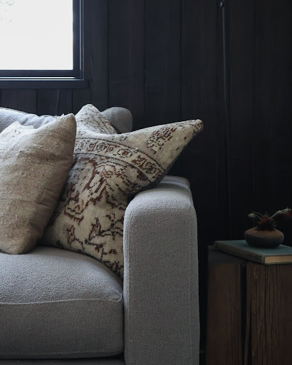 This image shows a living room featuring a dark wooden accent wall, a cozy light-colored couch with plush cushions, and a large window in the background allowing natural light to brighten the space.