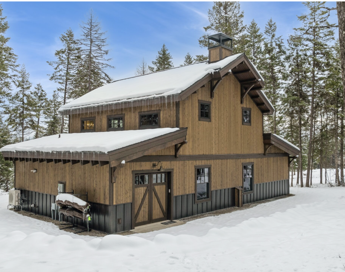 A modern two-story forest cabin has snow-covered surroundings and an exterior finished in thermally modified wood. 