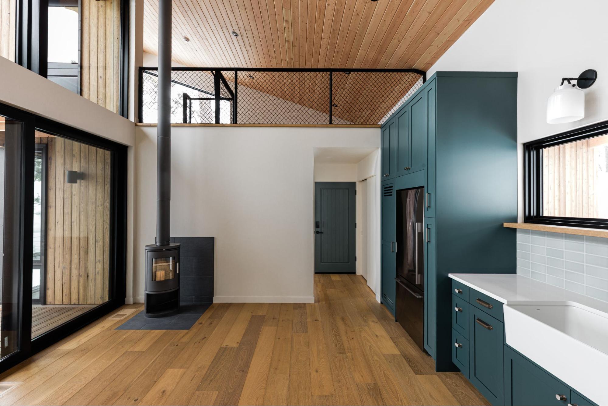 An open-concept interior features a warm-toned thermally modified wood ceiling complemented by teal cabinetry and black-framed windows. 
